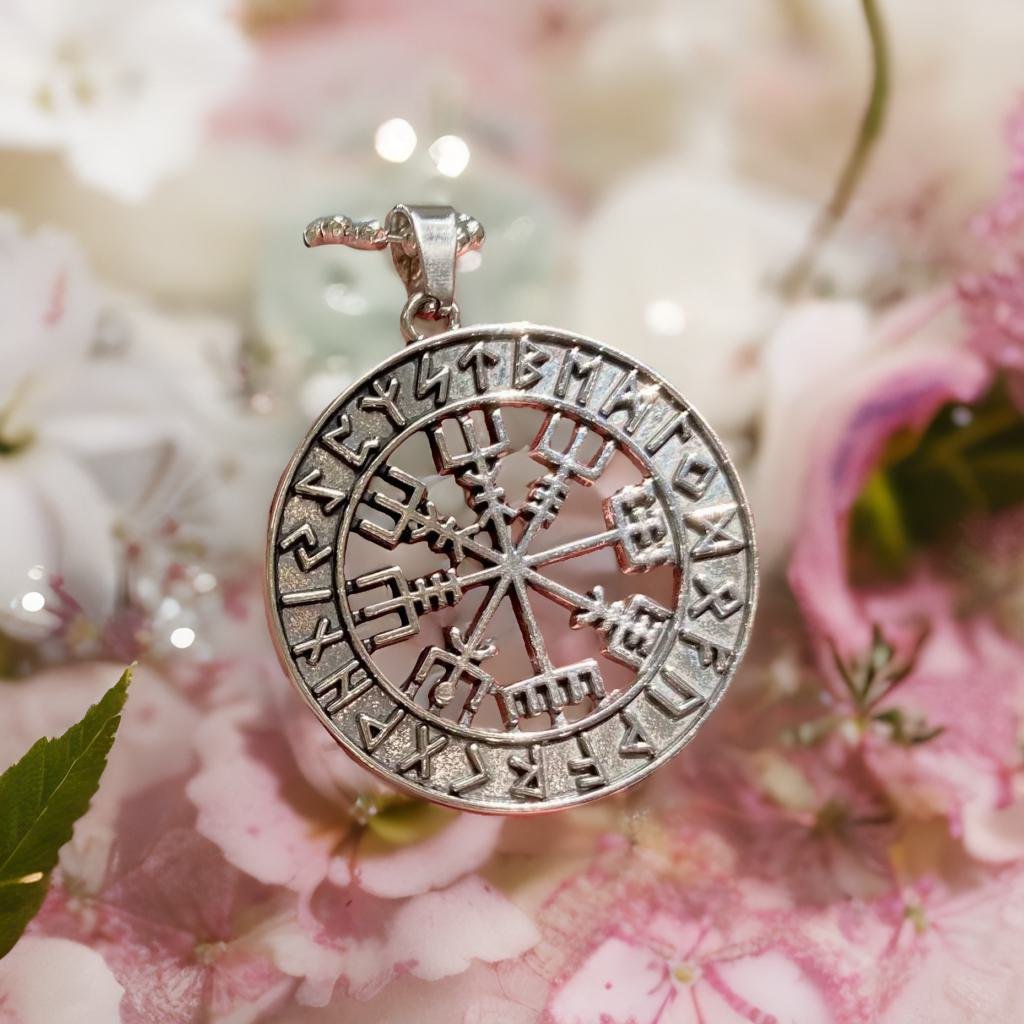 A silver pendant with a compass design is displayed against a background of pink and white flowers.