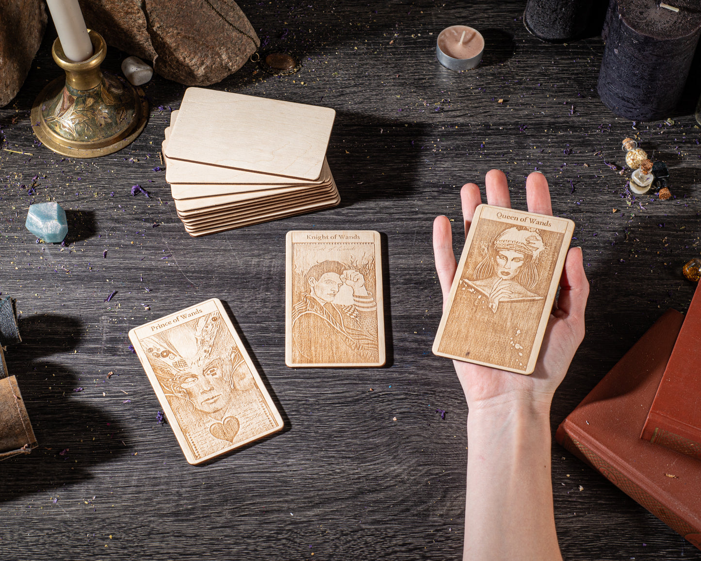 A hand is holding three wooden tarot cards on a dark wooden surface, surrounded by various objects such as candles, crystals, and books.
