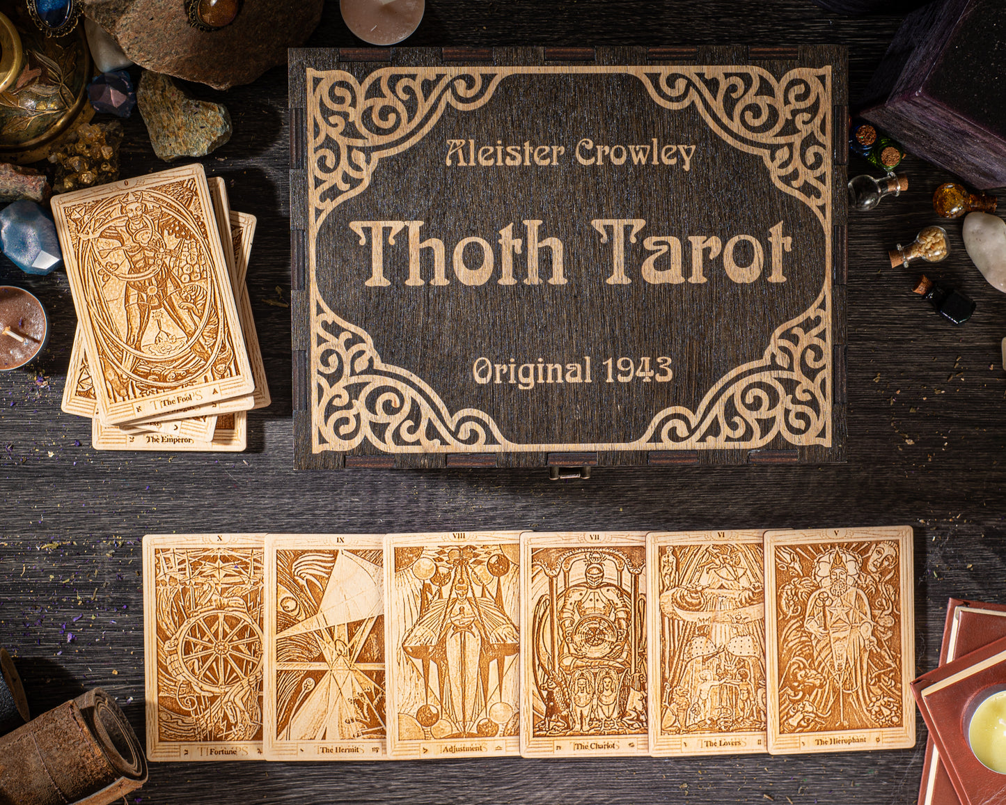 a collection of tarot cards and a wooden box with the name "Thoth Tarot" and the year "Original 1943" printed on it. The tarot cards are arranged on a dark wooden surface, and the wooden box is placed next to the cards.