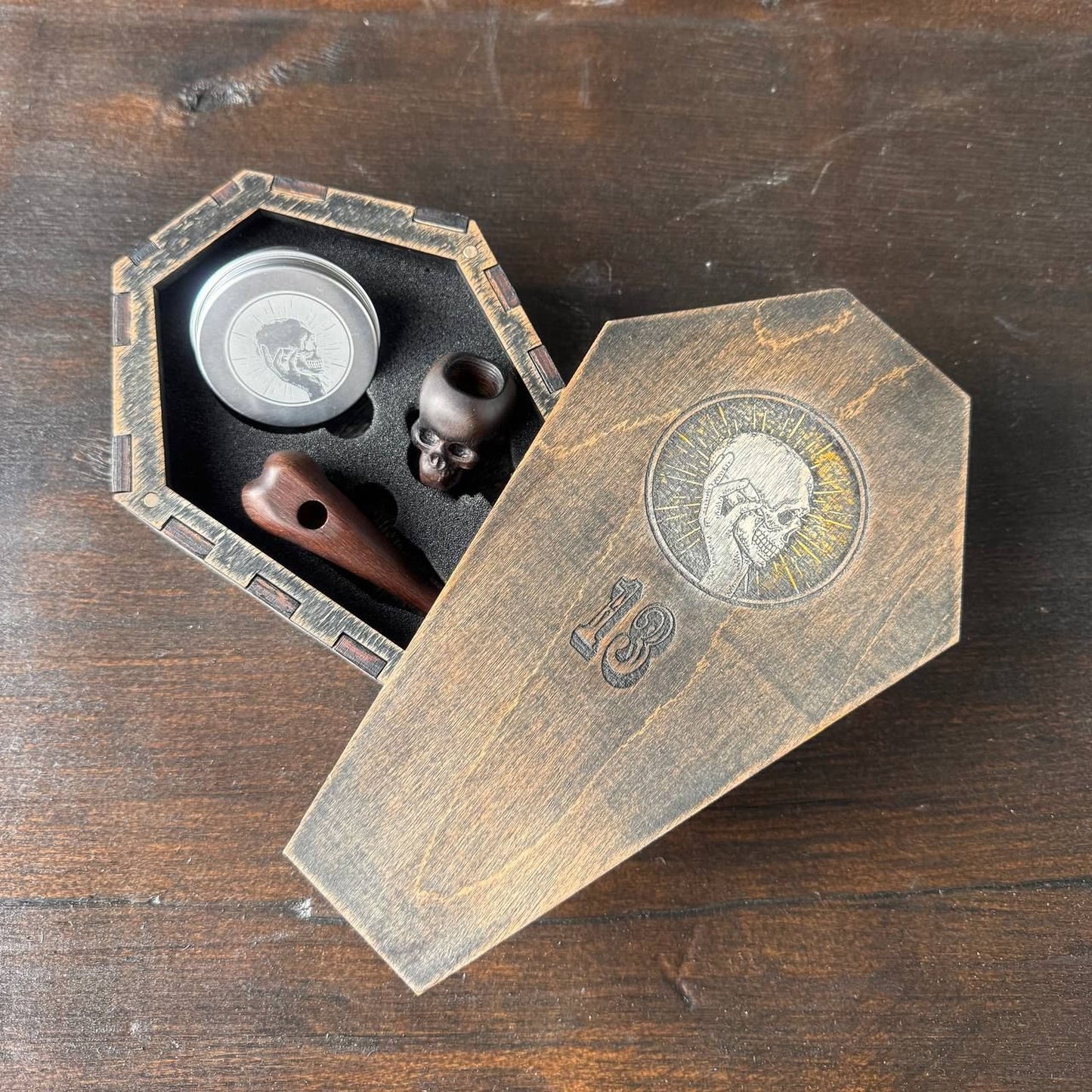 A wooden box with a skull design on it, containing a small wooden object and a silver object, placed on a wooden surface.