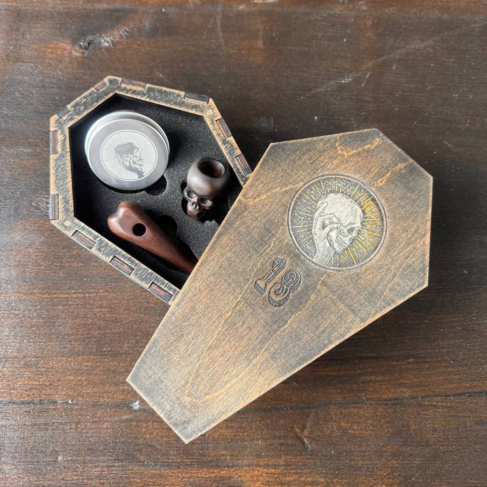 A wooden box with a skull design on it, containing a small wooden object and a silver object, placed on a wooden surface.
