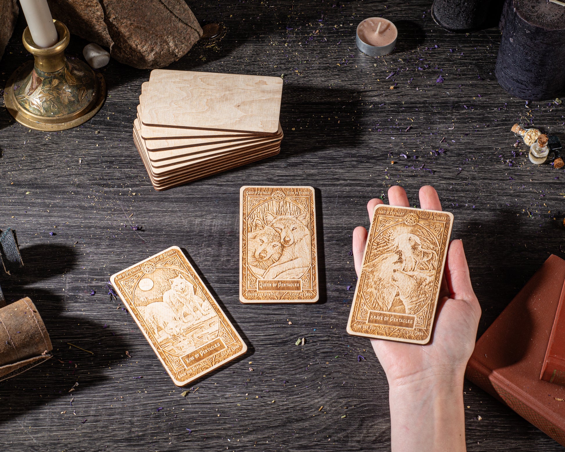 a hand holding three wooden tarot cards on a dark wooden surface, surrounded by various objects such as candles, books, and other items.