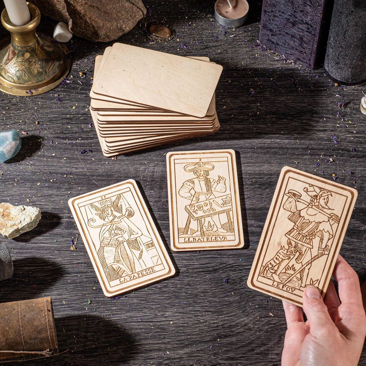 a hand holding three wooden tarot cards, each with a unique illustration and text, placed on a wooden surface.