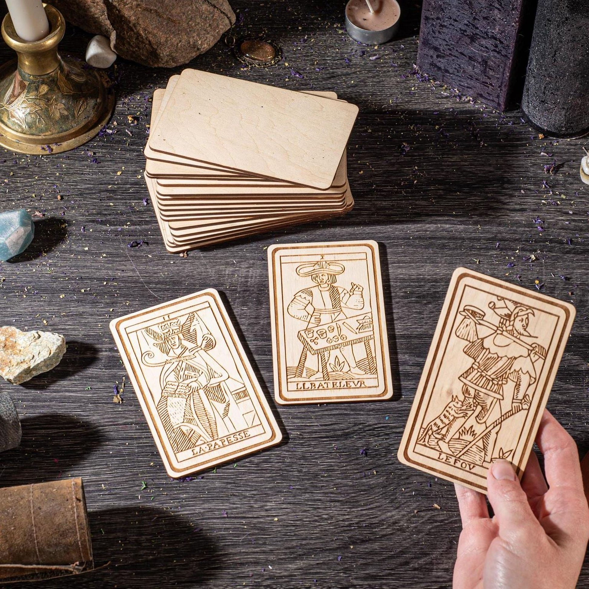 a hand holding three wooden tarot cards, each with a unique illustration and text, placed on a wooden surface.