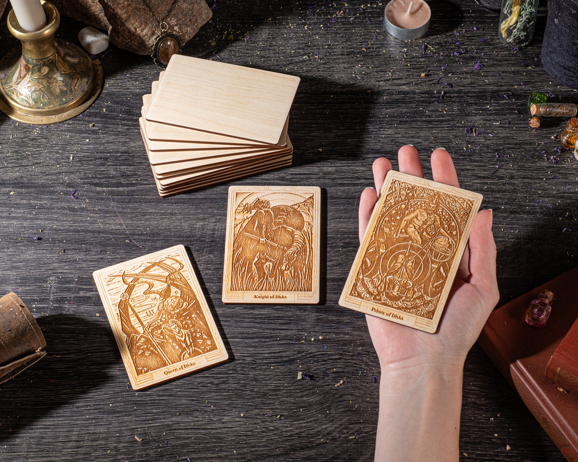 A hand is holding three wooden tarot cards on a dark wooden surface, surrounded by various objects such as candles, crystals, and other items.