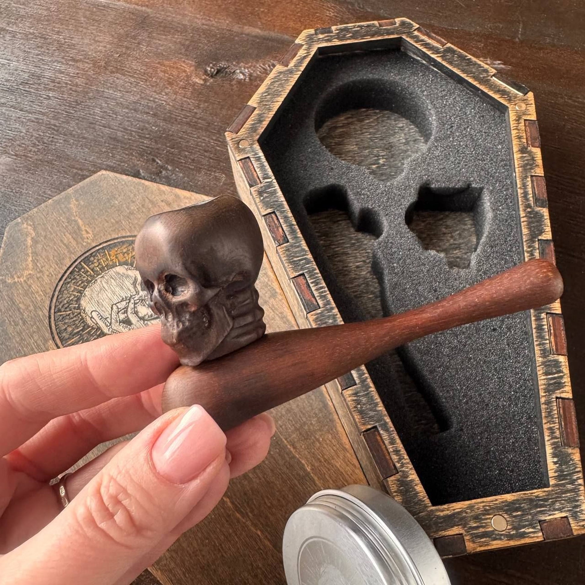 A hand holding a wooden pipe with a skull-shaped handle, placed next to a wooden box with a carved lid.