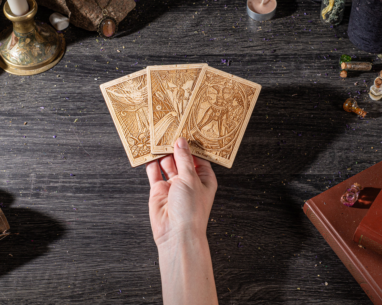 A hand holding a deck of tarot cards on a wooden surface, surrounded by various objects such as a candle, books, and crystals.