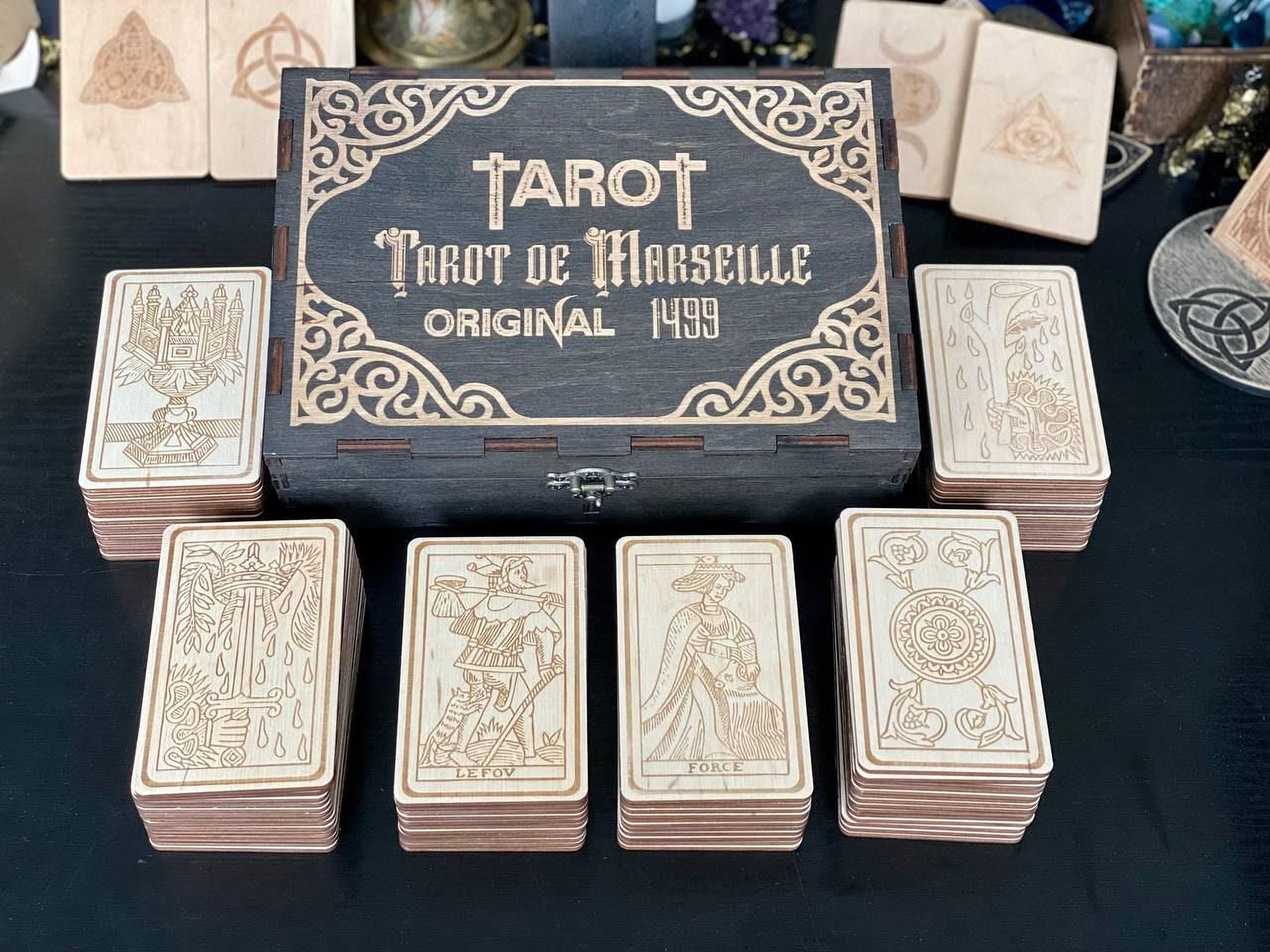 a collection of tarot cards and a wooden box with the text "Tarot de Marseille" on it. The cards are arranged in stacks on a table, and the box is placed in the center of the image.