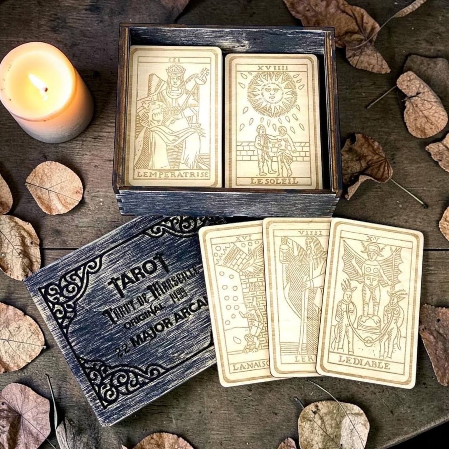 a wooden box containing four tarot cards, a lit candle, and some fallen leaves on a wooden surface.