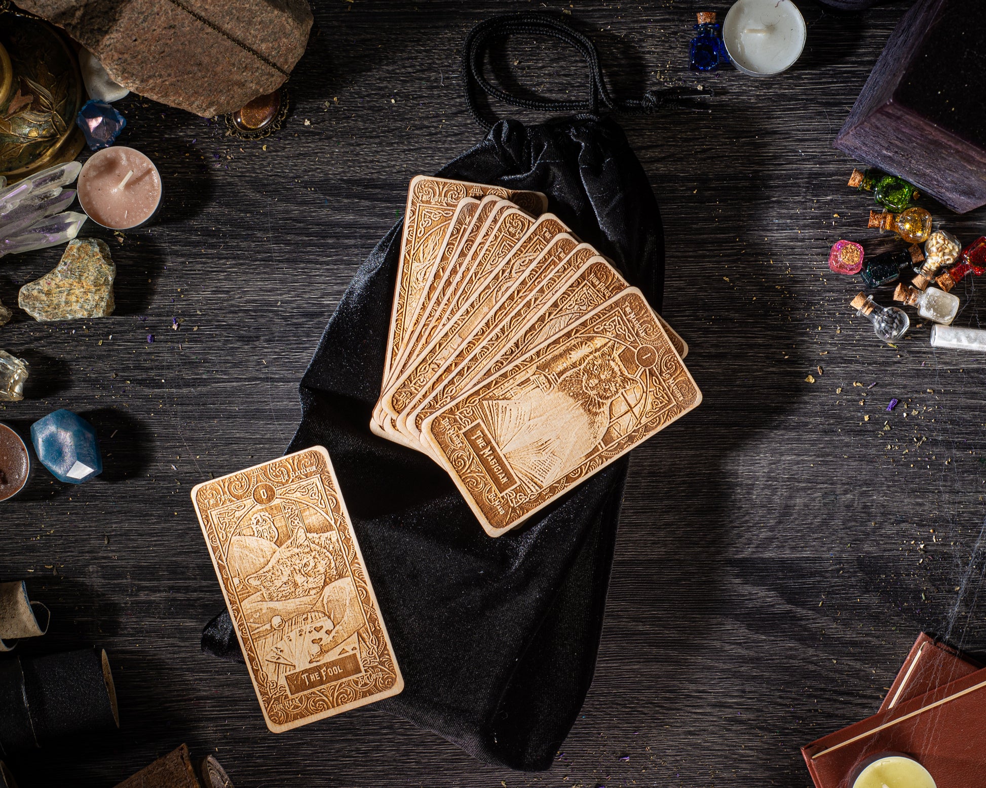 A collection of wooden tarot cards, a black velvet pouch, and various other items such as crystals, candles, and books are arranged on a dark wooden surface.