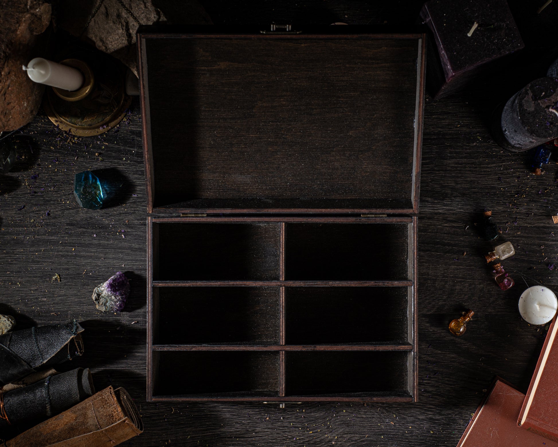 An open wooden box with empty compartments sits on a dark wooden surface, surrounded by various objects such as candles, gemstones, and books.