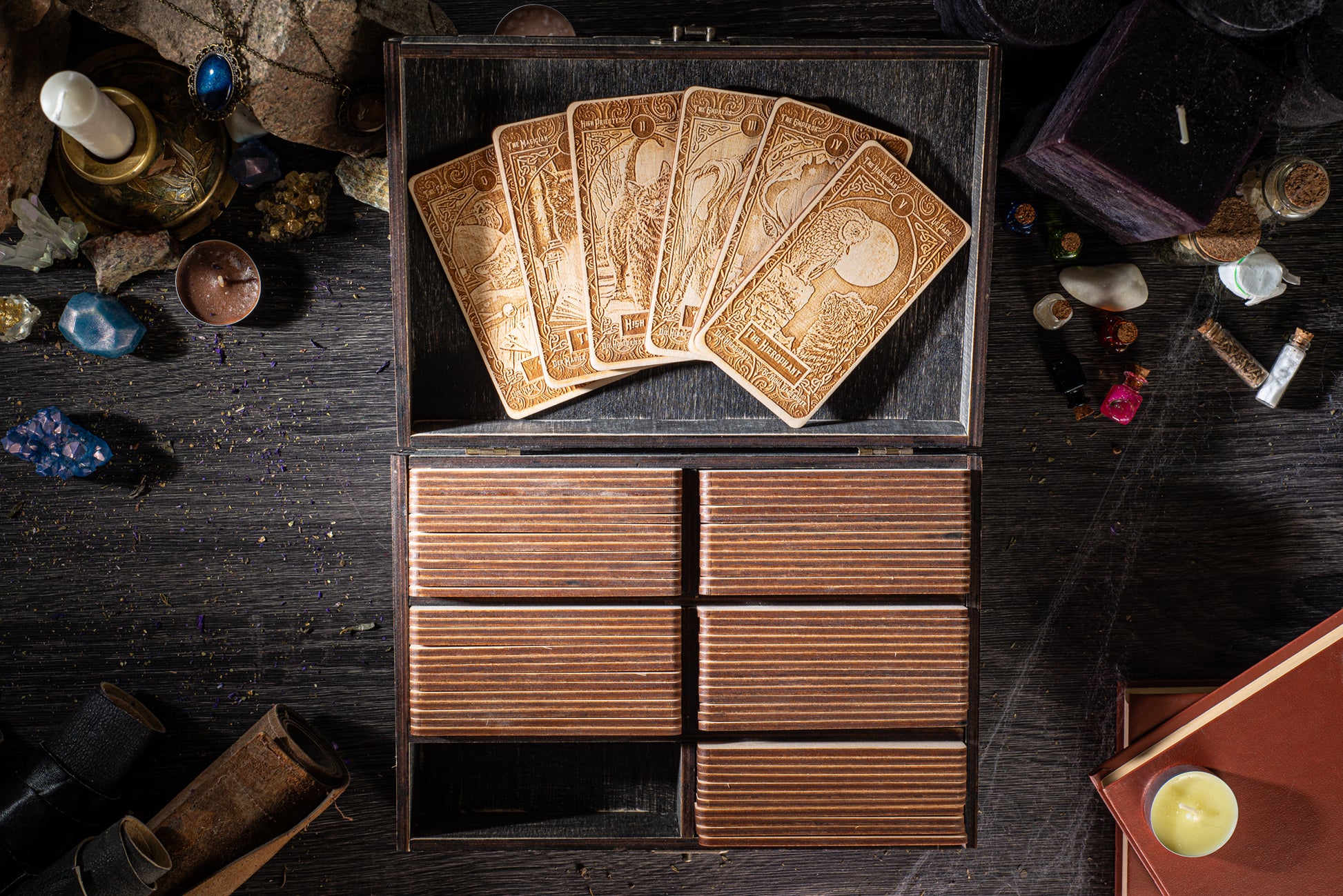 a wooden box with six compartments, containing a deck of playing cards, and various other items such as crystals, candles, and books scattered around the box on a dark wooden surface.