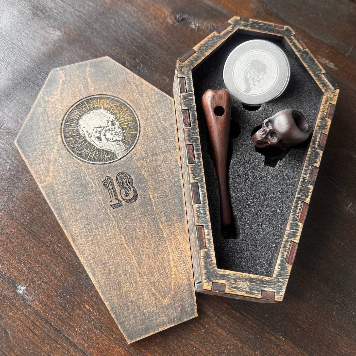 A wooden box with a skull design and the number 18 on it, along with a wooden pipe and a small wooden object inside the box.