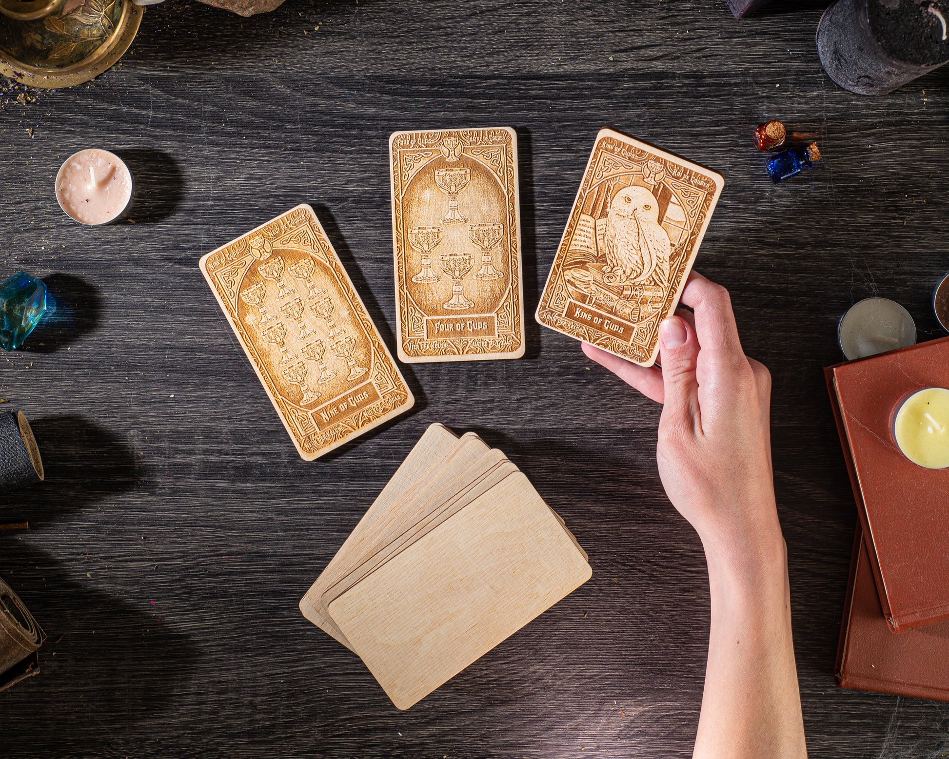 a hand holding three wooden tarot cards on a dark wooden surface, surrounded by various objects such as candles, books, and other items.