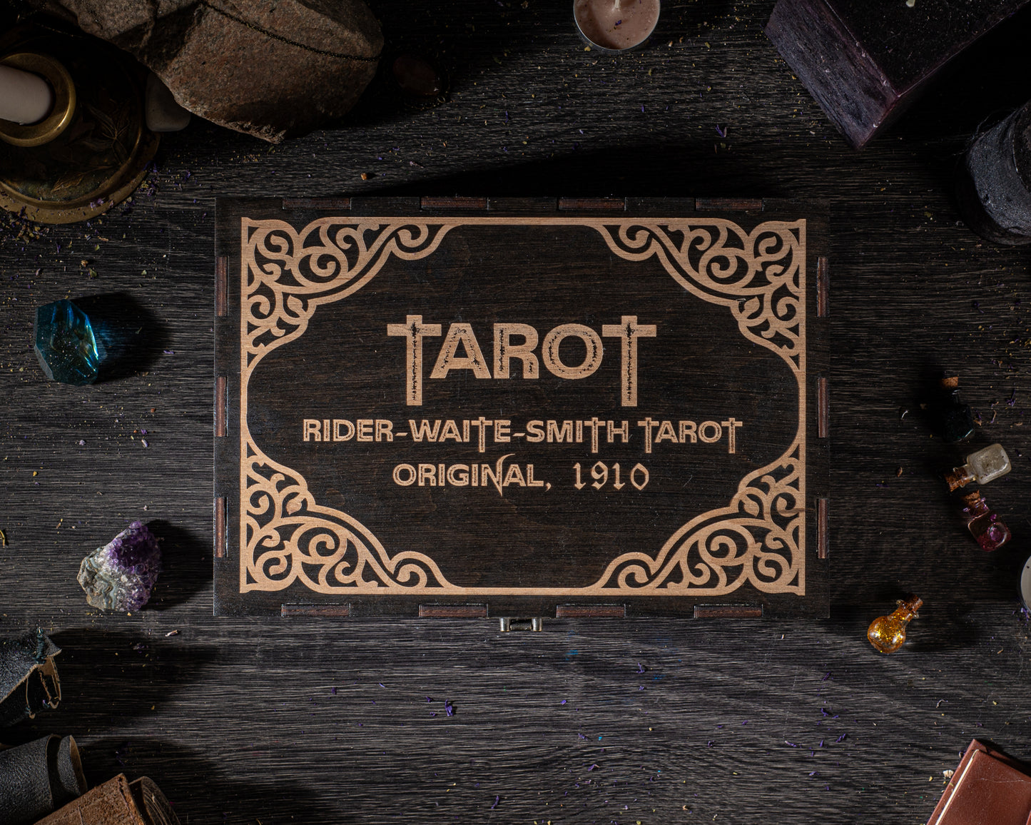 A wooden box with the name 'TAROT' and the text 'RIDER-WAITE-SMITH TAROT ORIGINAL, 1910' engraved on it, surrounded by various objects such as crystals, bottles, and a candle on a dark wooden surface.