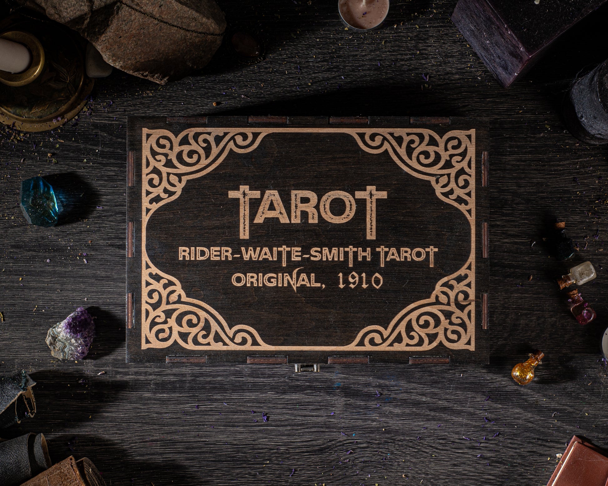 A wooden box with the name 'TAROT' and the text 'RIDER-WAITE-SMITH TAROT ORIGINAL, 1910' engraved on it, surrounded by various objects such as crystals, bottles, and a candle on a dark wooden surface.