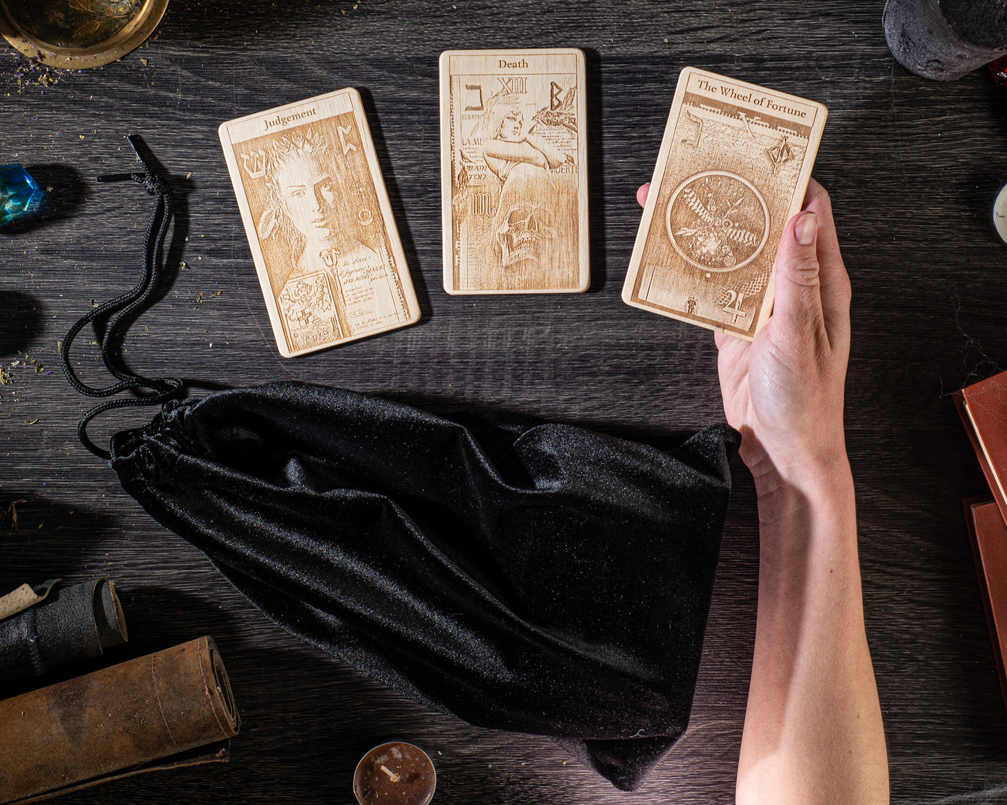 a person's hand holding three wooden tarot cards, each with a different design, placed on a wooden surface alongside a black velvet pouch and some other items.