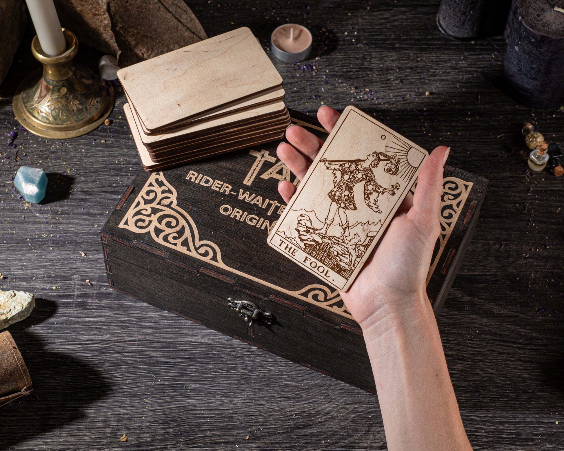 A hand holding a wooden tarot card with a design on it, placed on a wooden box with the text "RIDER-WAITER ORIGINAL THE FOIL" printed on it.