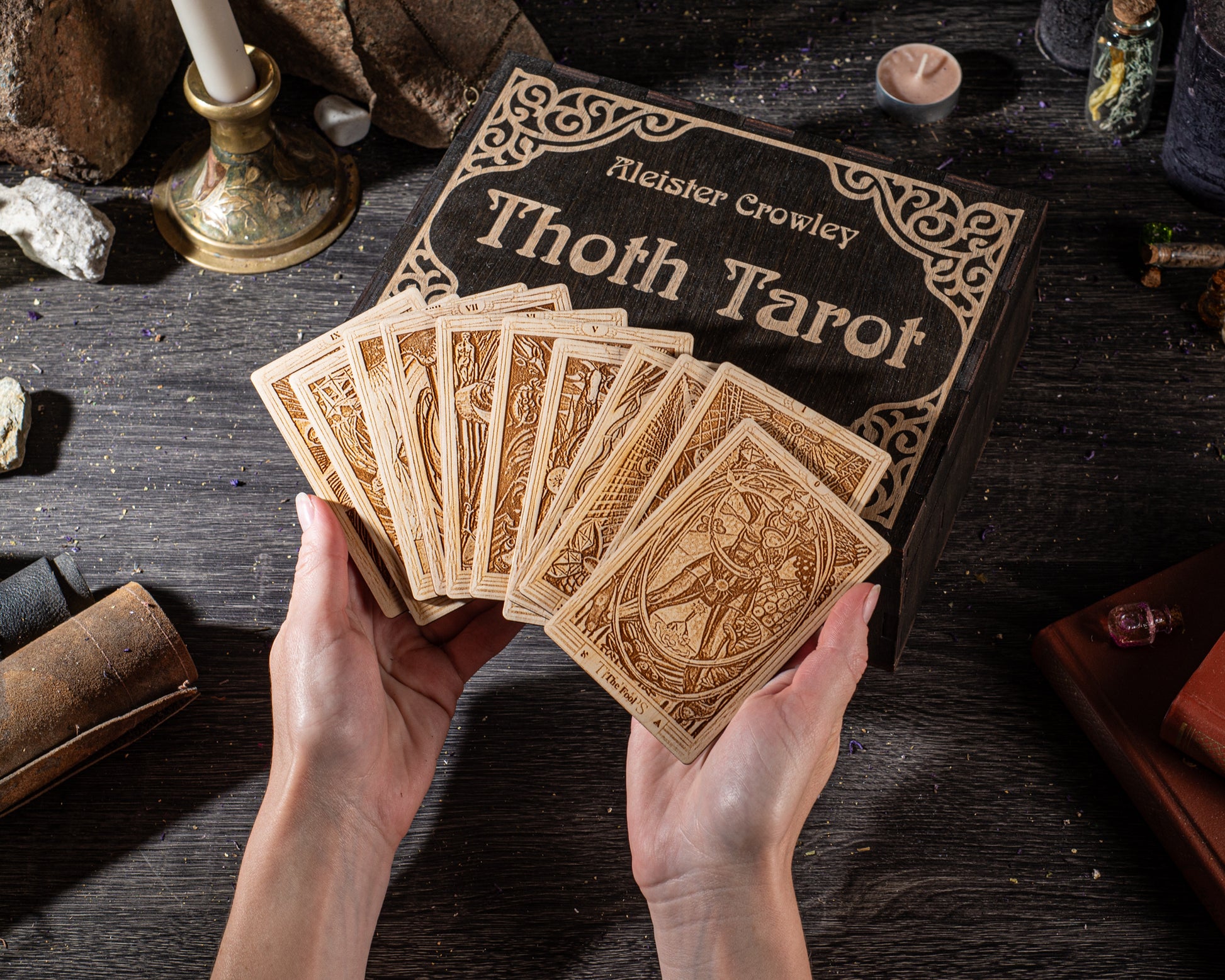 A person's hands holding a deck of cards with a book titled "Thoth Tarot" in the background.