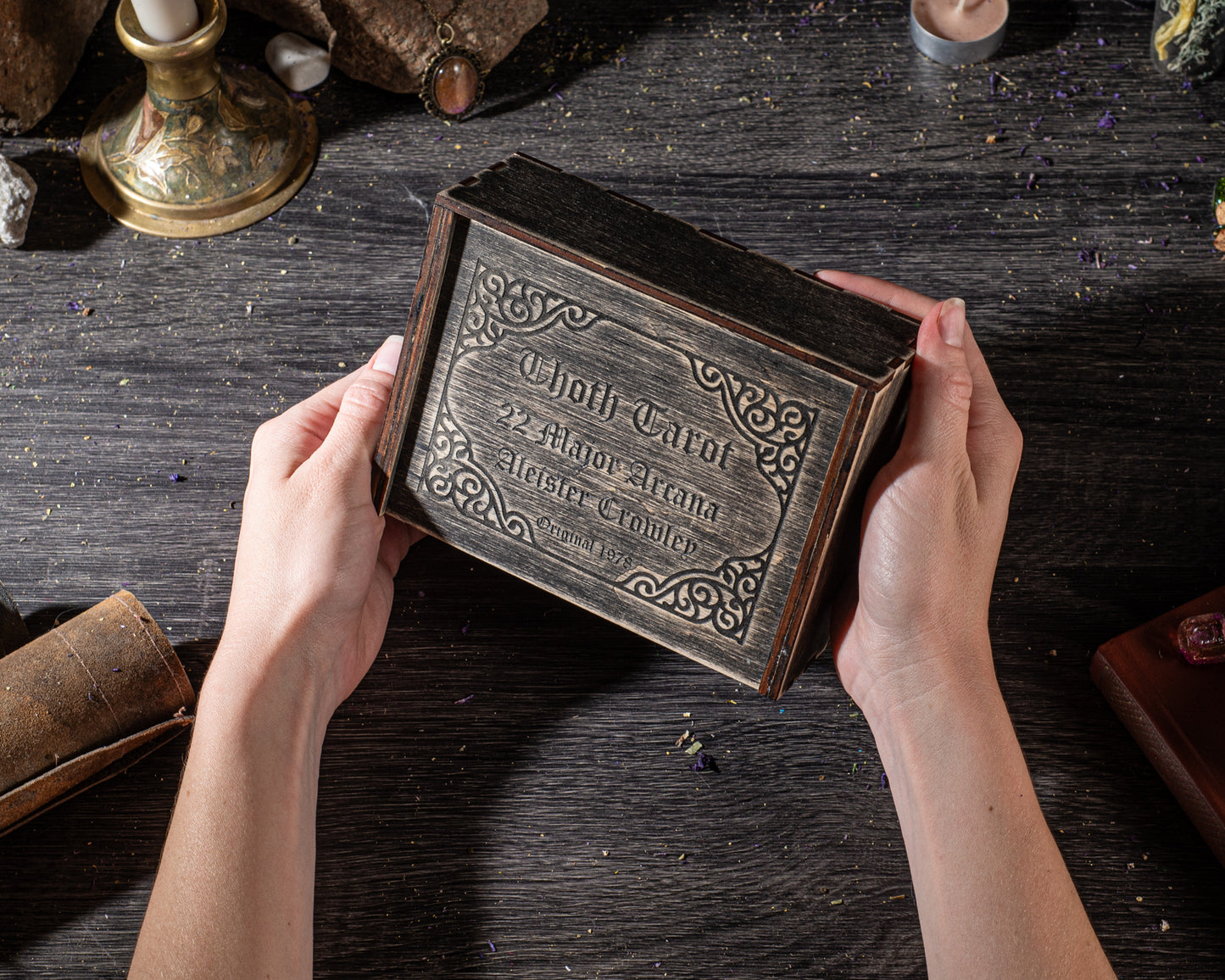 A person's hands holding a wooden box with an ornate design, placed on a wooden surface.