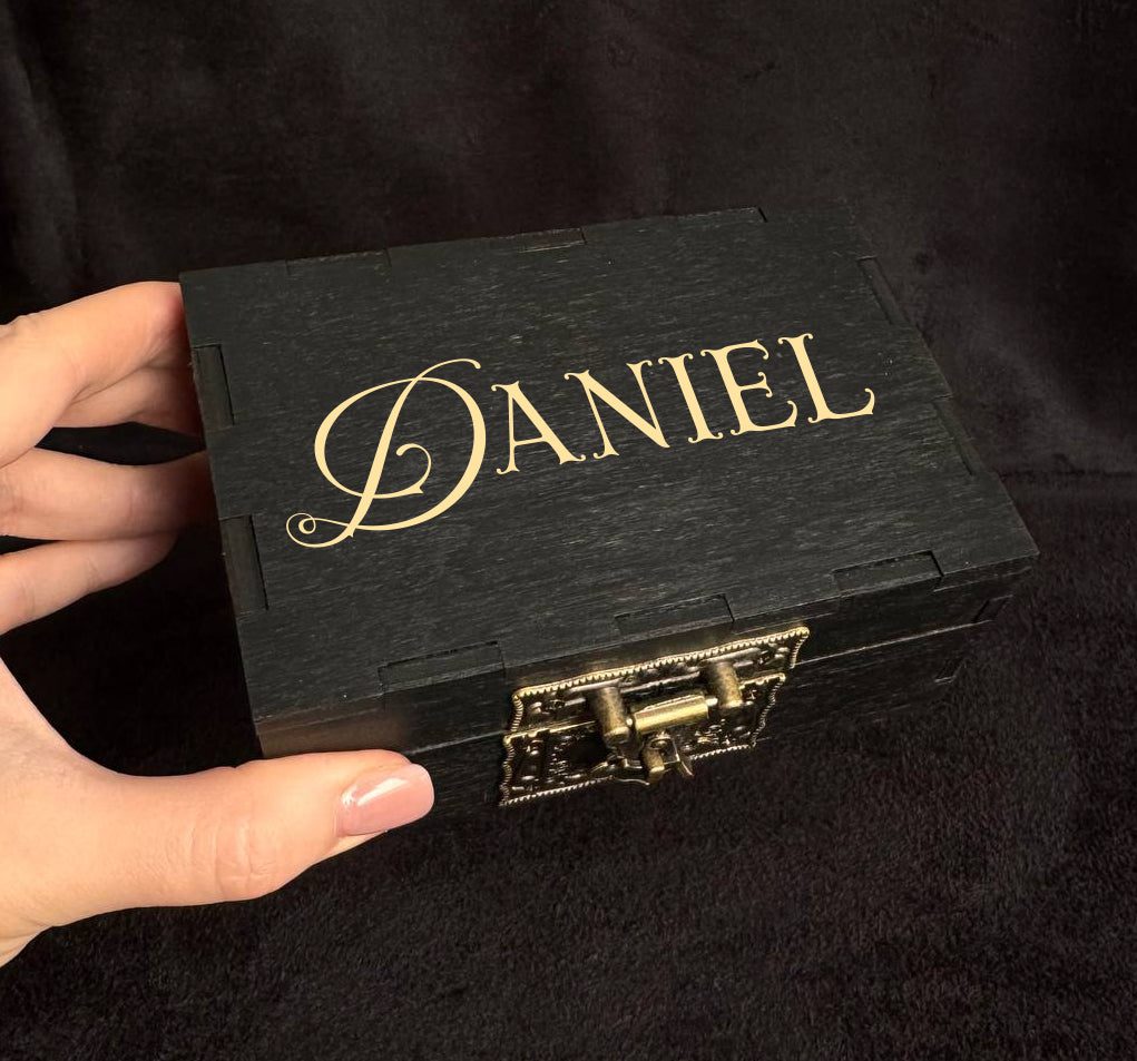 A hand is holding a black wooden box with the name "DANIEL" engraved on it.