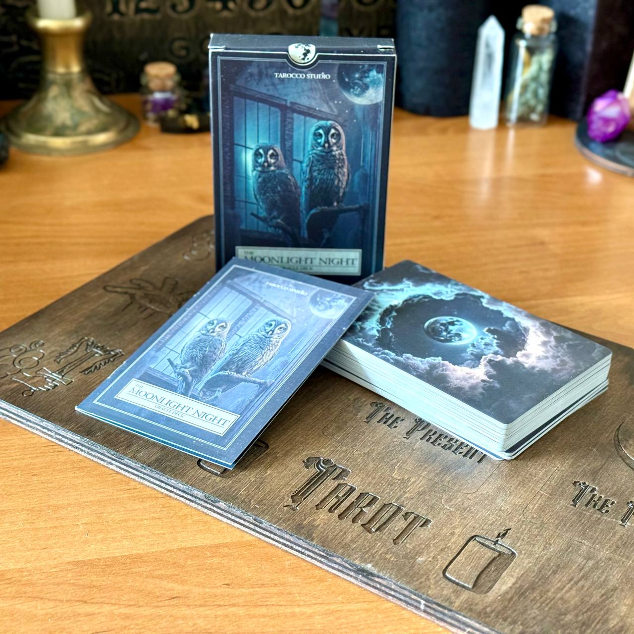a tarot deck with a wooden board displaying the words "Moonlight Night" and "The Present" on it. The deck includes a book and a card set, with the book lying on top of the wooden board and the card set placed on the board.