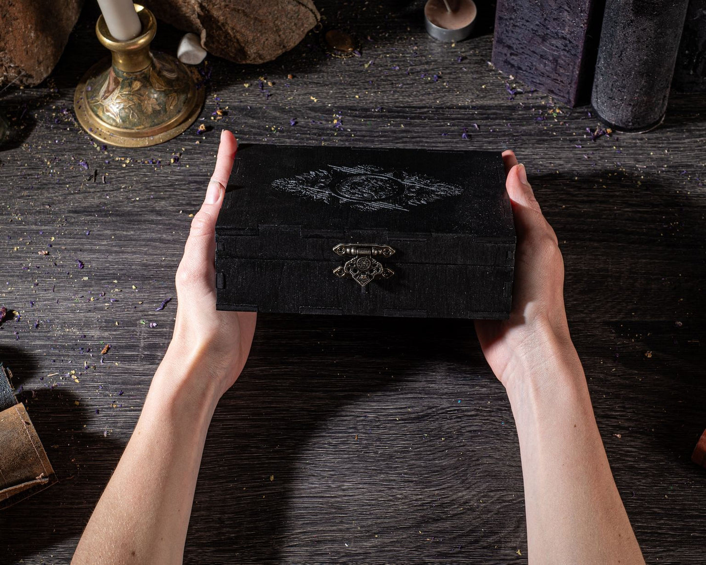 A pair of hands holding a black wooden box with a decorative design on it, placed on a wooden surface with various objects in the background.