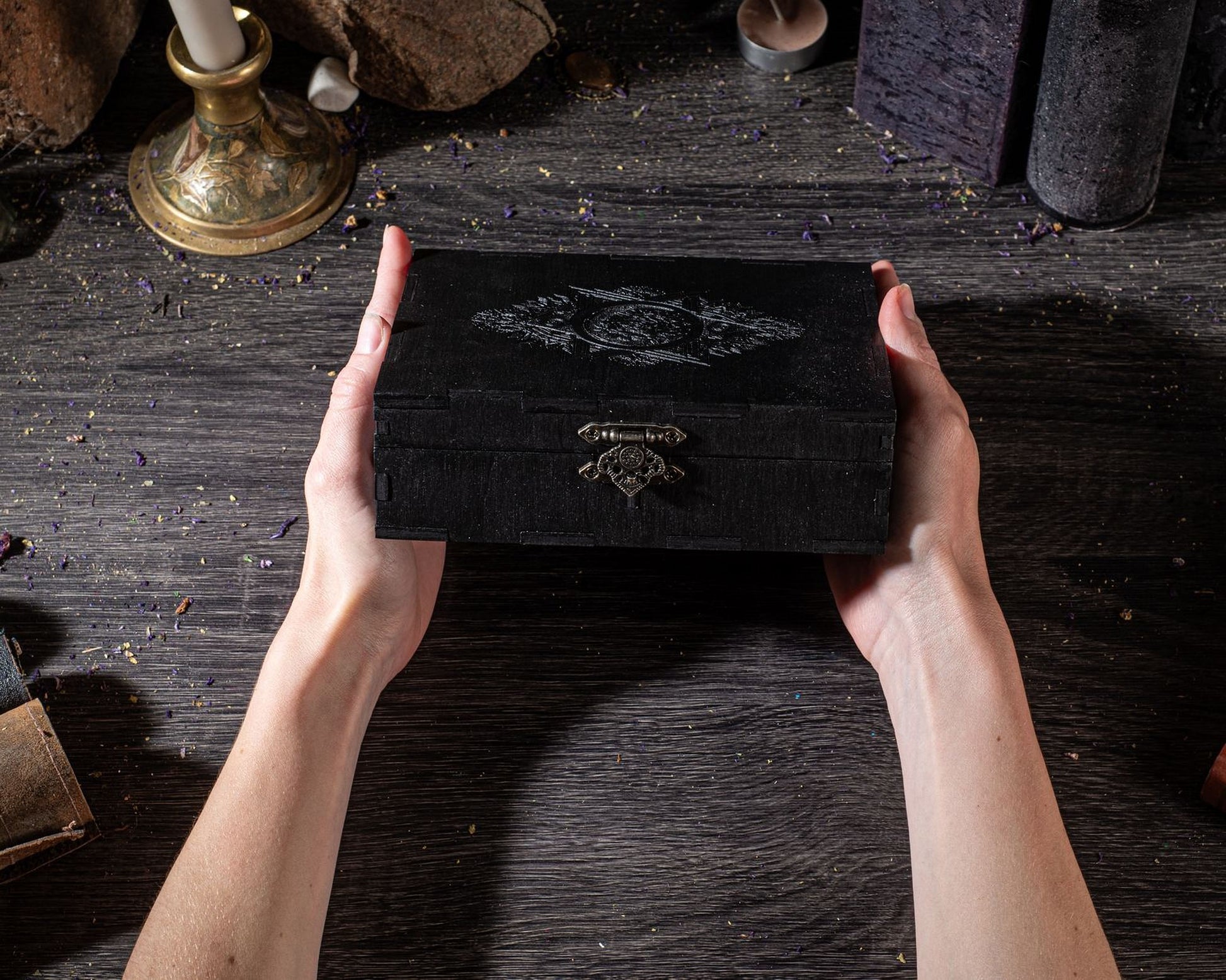 A pair of hands holding a black wooden box with a decorative design on it, placed on a wooden surface with various objects in the background.