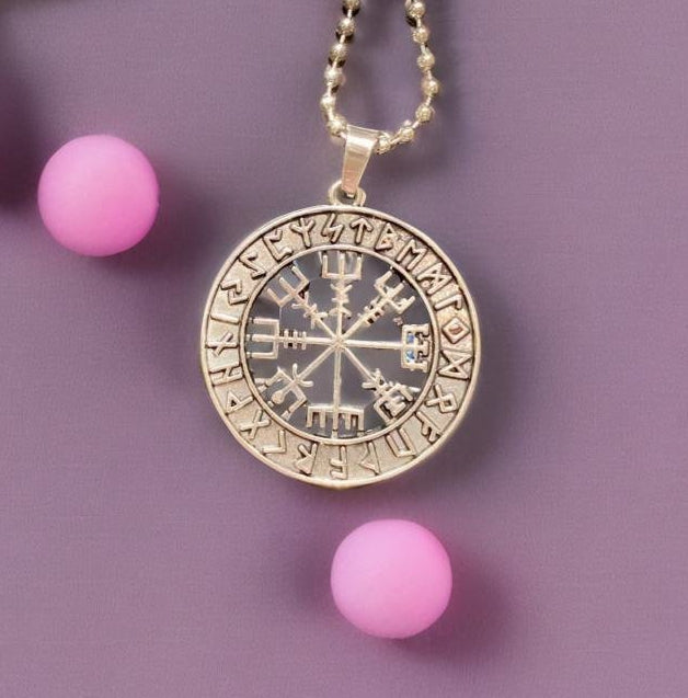 A gold-colored pendant necklace with a compass design, featuring a central compass with various symbols and numbers surrounding it, is displayed against a purple background.