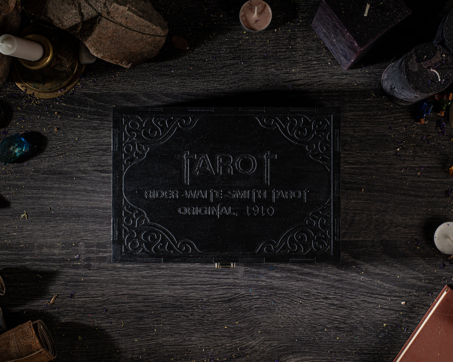 a black box with the word "TAROT" embossed on it, surrounded by various objects such as candles, gemstones, and other items on a wooden surface.