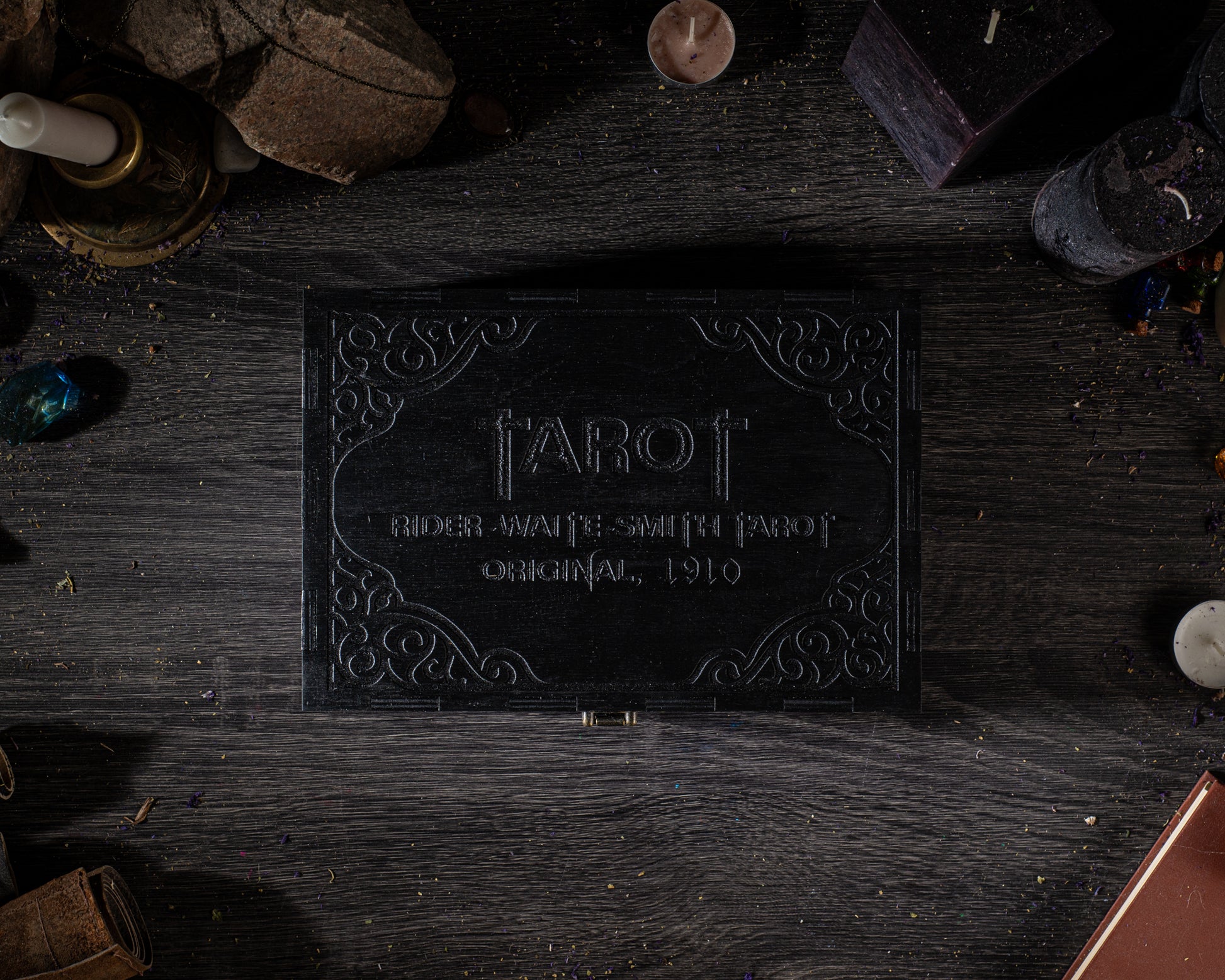a black box with the word "TAROT" embossed on it, surrounded by various objects such as candles, gemstones, and other items on a wooden surface.