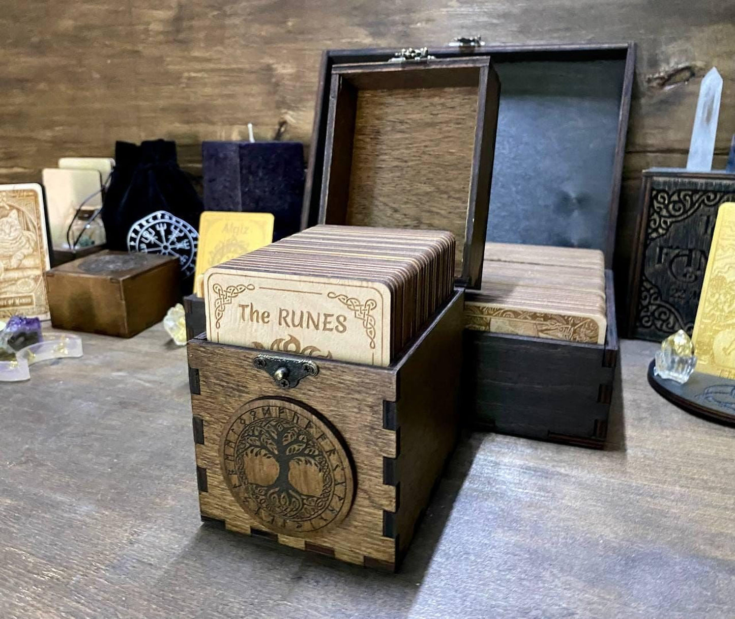a wooden box with a tree design on it, containing several cards with the text "The Ruins" printed on them. The box is placed on a wooden surface, and there are other items in the background, including a wooden box and a wooden crate.