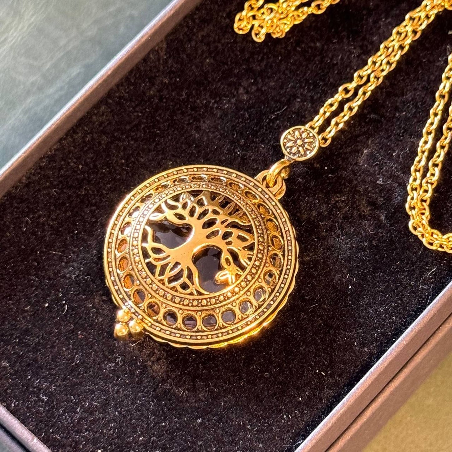 A gold-colored necklace with a circular pendant featuring a tree design, displayed in a dark box.