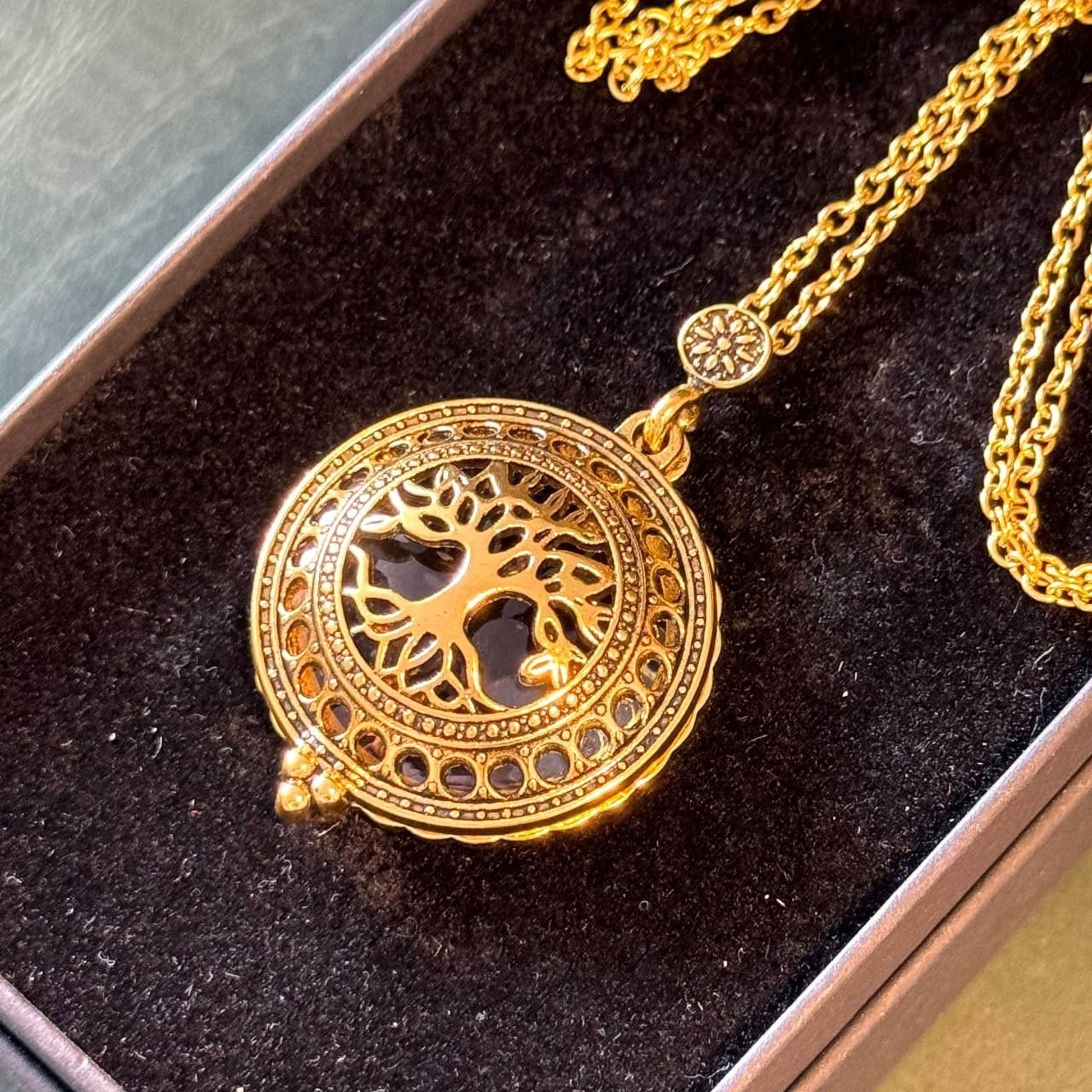 A gold-colored necklace with a circular pendant featuring a tree design, displayed in a dark box.