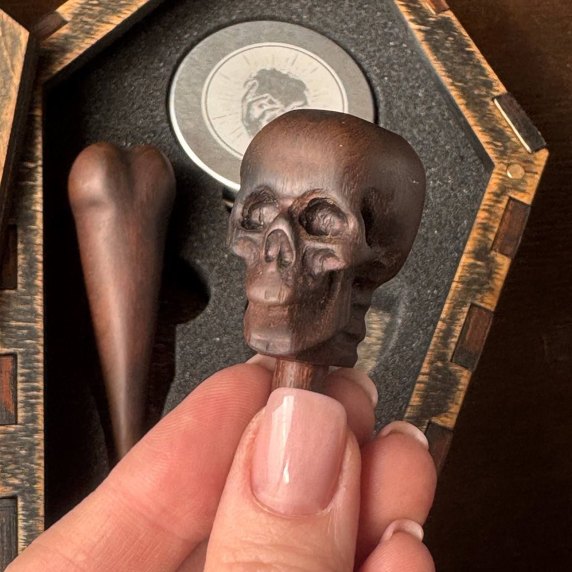 A hand holding a wooden skull-shaped object, which appears to be a pen or inkwell cap, against a dark background.