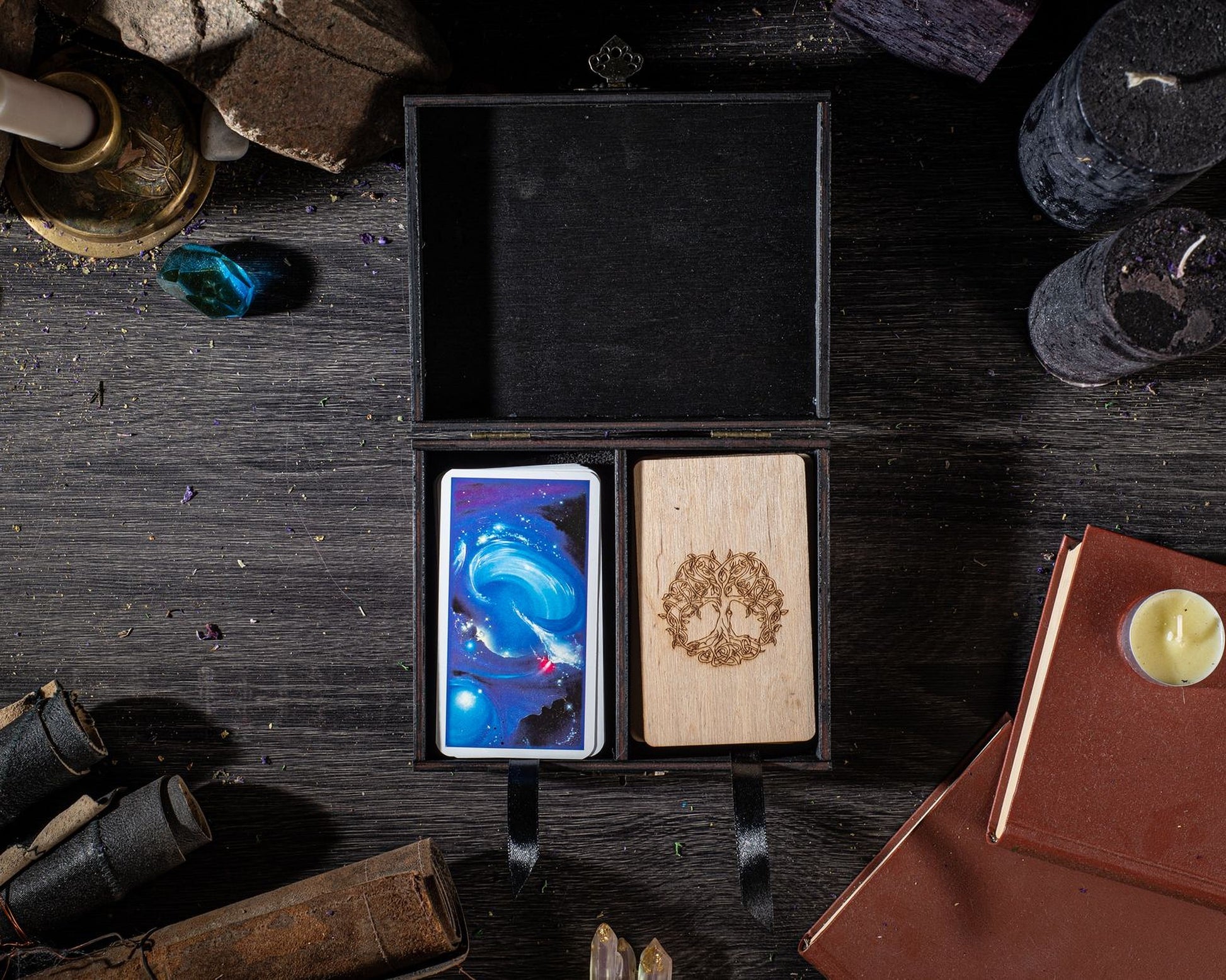 a wooden box with two cards inside, one of which has a tree design, placed on a dark wooden surface alongside various objects such as candles, books, and crystals.