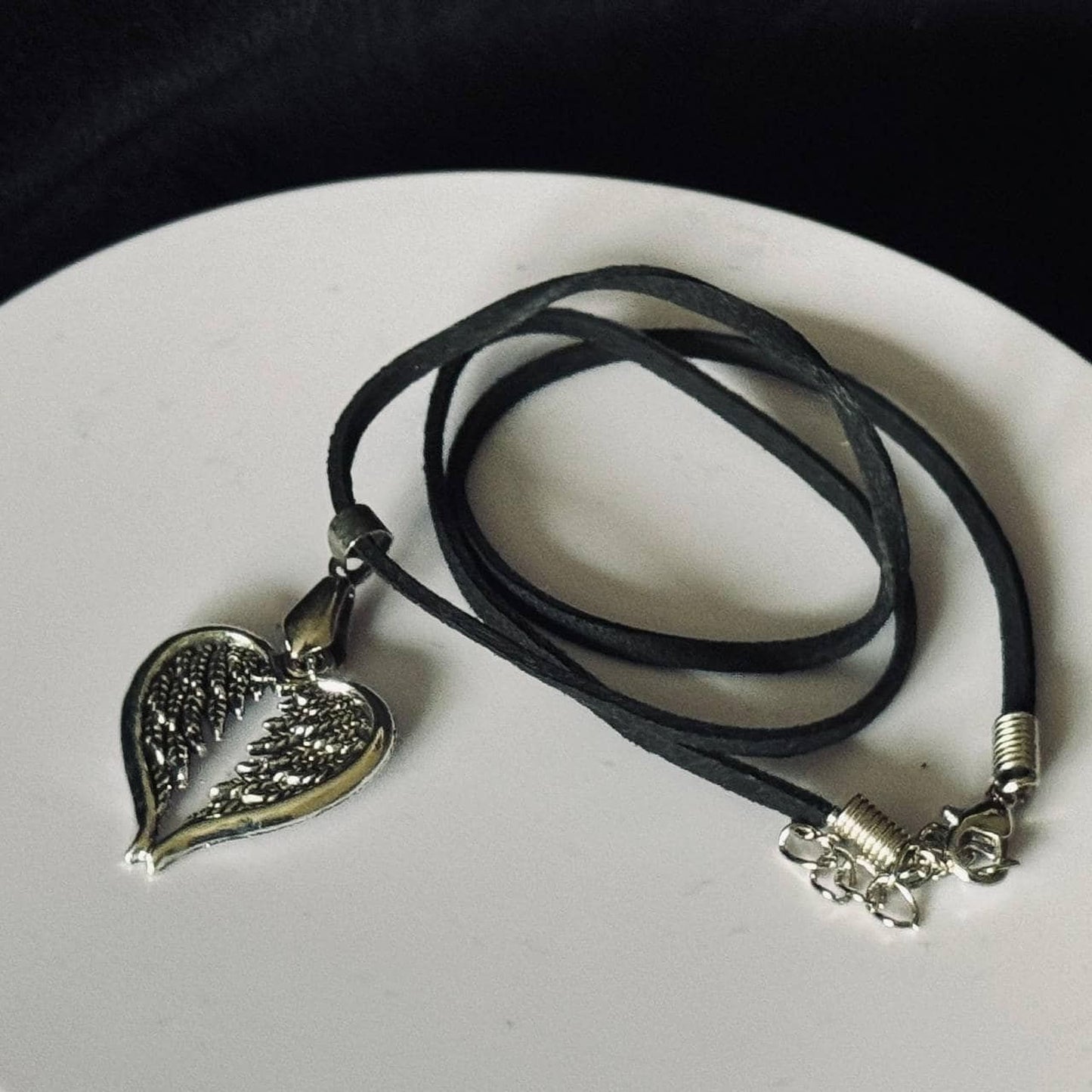 A silver heart-shaped pendant with wings is displayed on a black cord necklace.
