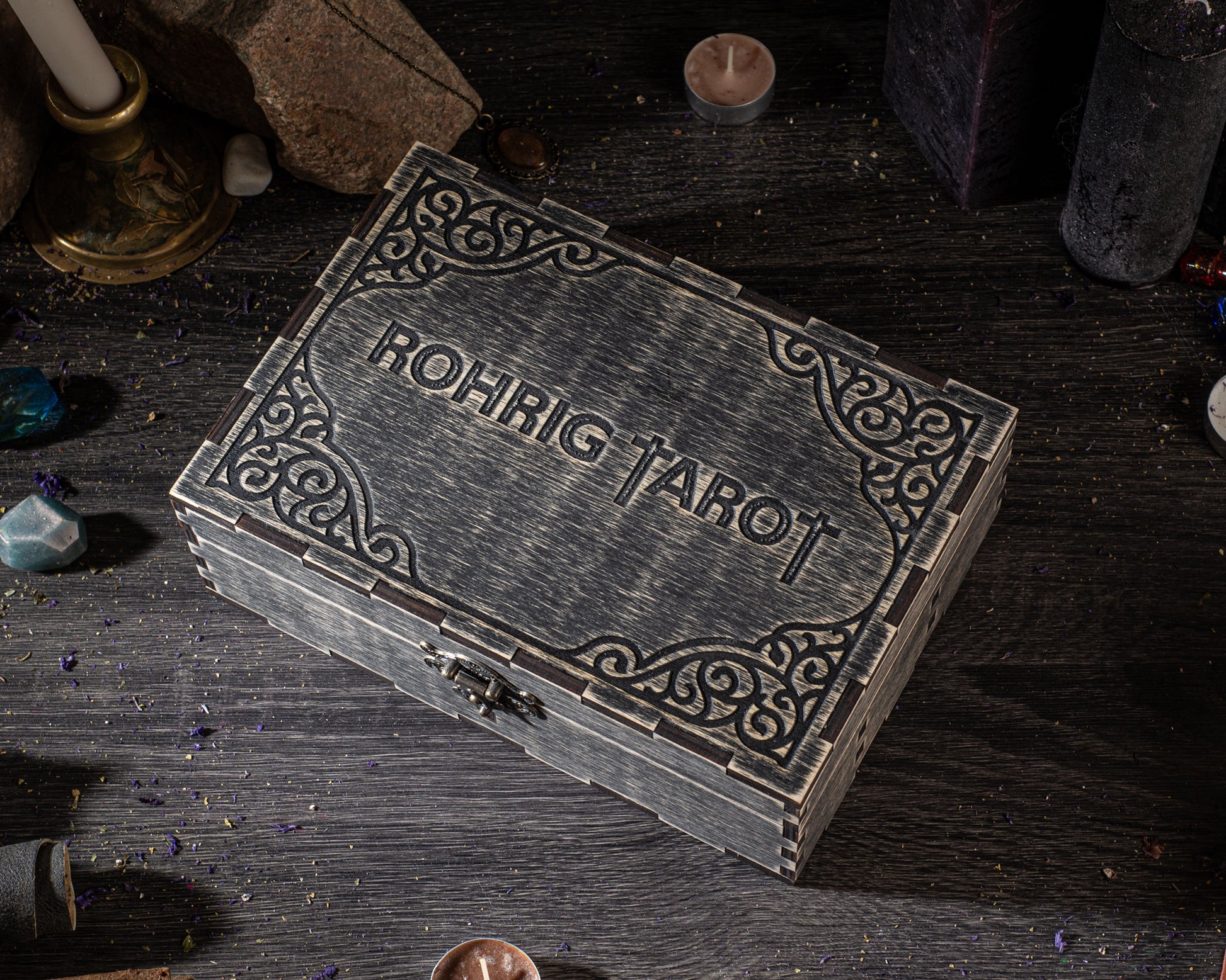 A wooden box with the name "Rohrig Tarot" embossed on it, surrounded by various objects such as candles, gemstones, and other items on a dark wooden surface.