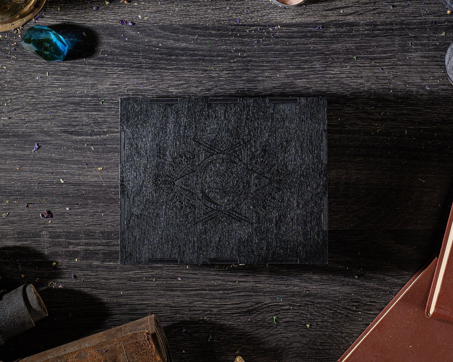 a black square box with a carved design on it, placed on a wooden surface with scattered objects around it.