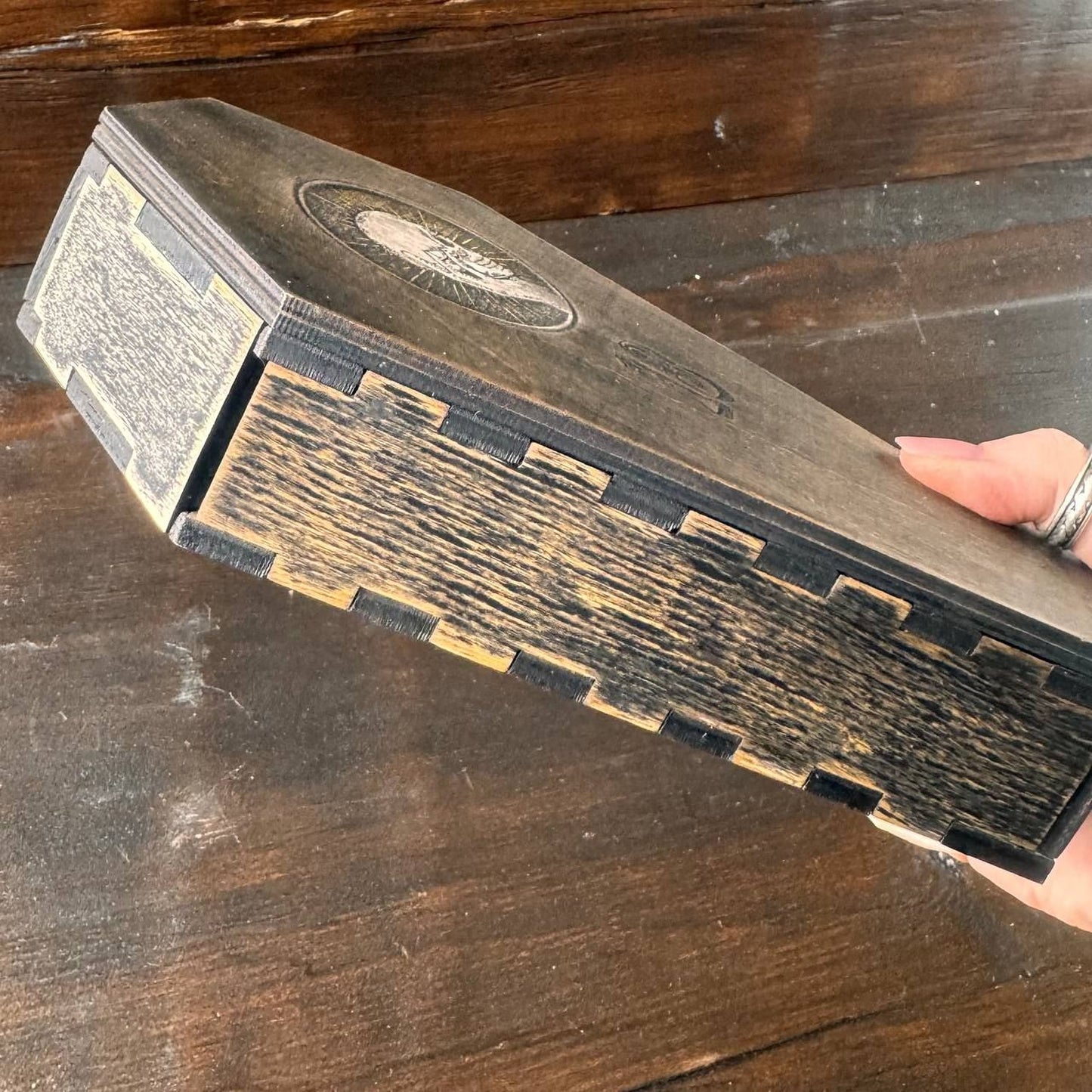 A hand is holding a wooden box with a carved design on the top, resting on a wooden surface.
