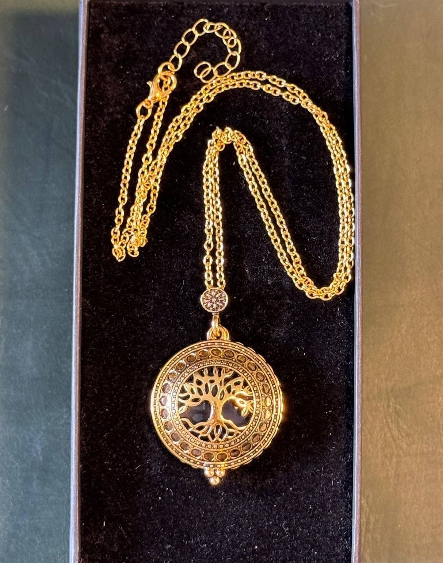 A gold-colored necklace with a circular pendant featuring a tree design, displayed in a black velvet box.