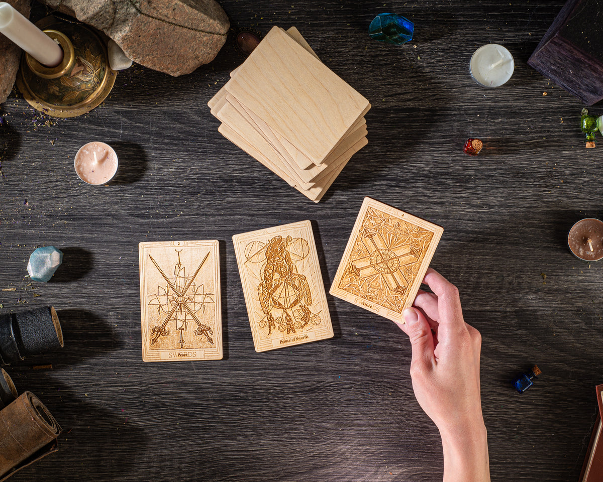 a hand holding a wooden card with intricate designs, surrounded by various objects such as candles, crystals, and other items on a dark wooden surface.