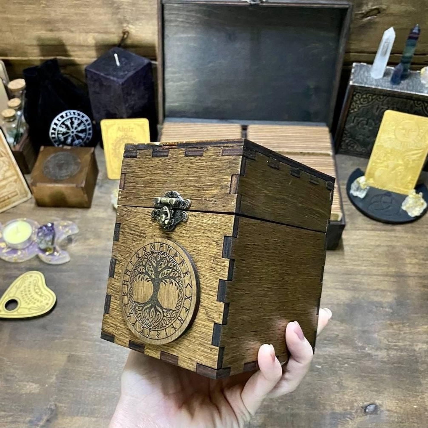 A hand is holding a wooden box with a tree design on it, surrounded by various objects on a wooden surface.