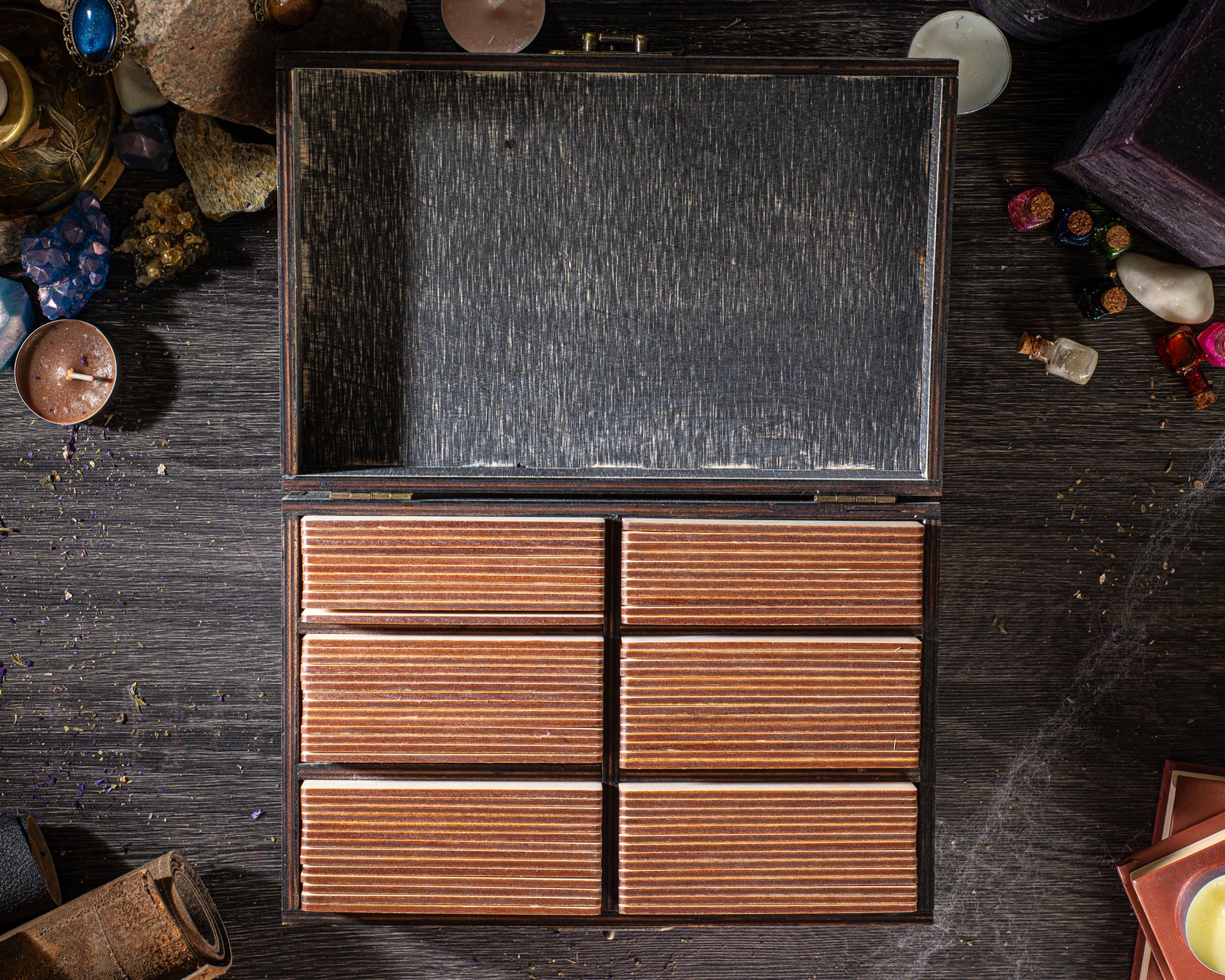 an open wooden chest with six compartments, placed on a wooden surface. The chest appears to be made of wood and has a dark, textured finish. There are various objects scattered around the chest, including a small white object, a few books, and some other items. The overall scene suggests a collection or display of various objects.