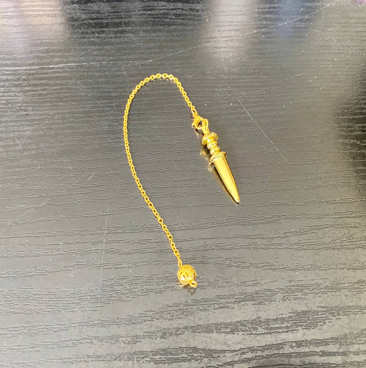 A gold-colored pendant with a small ball at the end is shown on a wooden surface.
