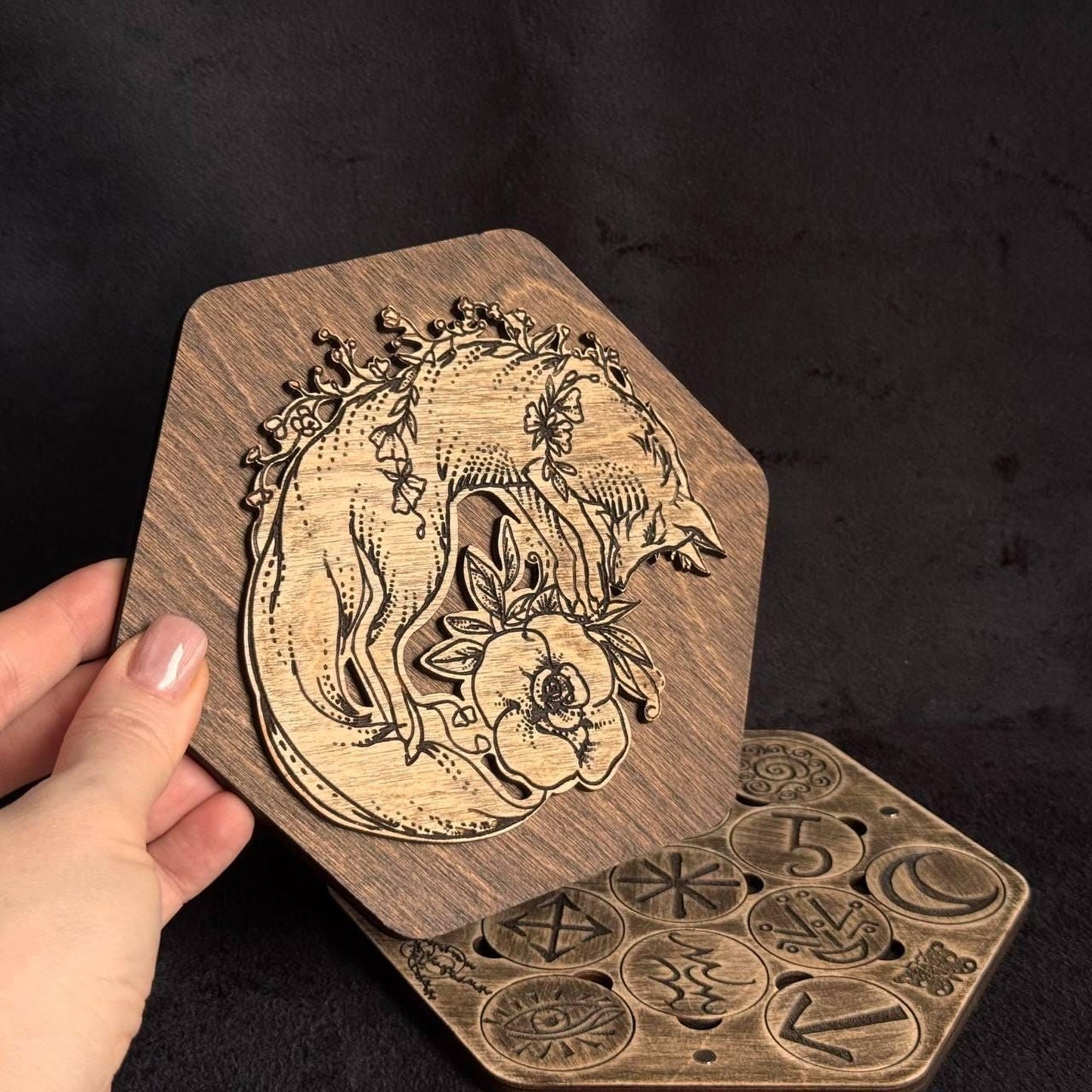 A hand is holding a wooden object with an intricate engraved design, including a fox and floral elements.