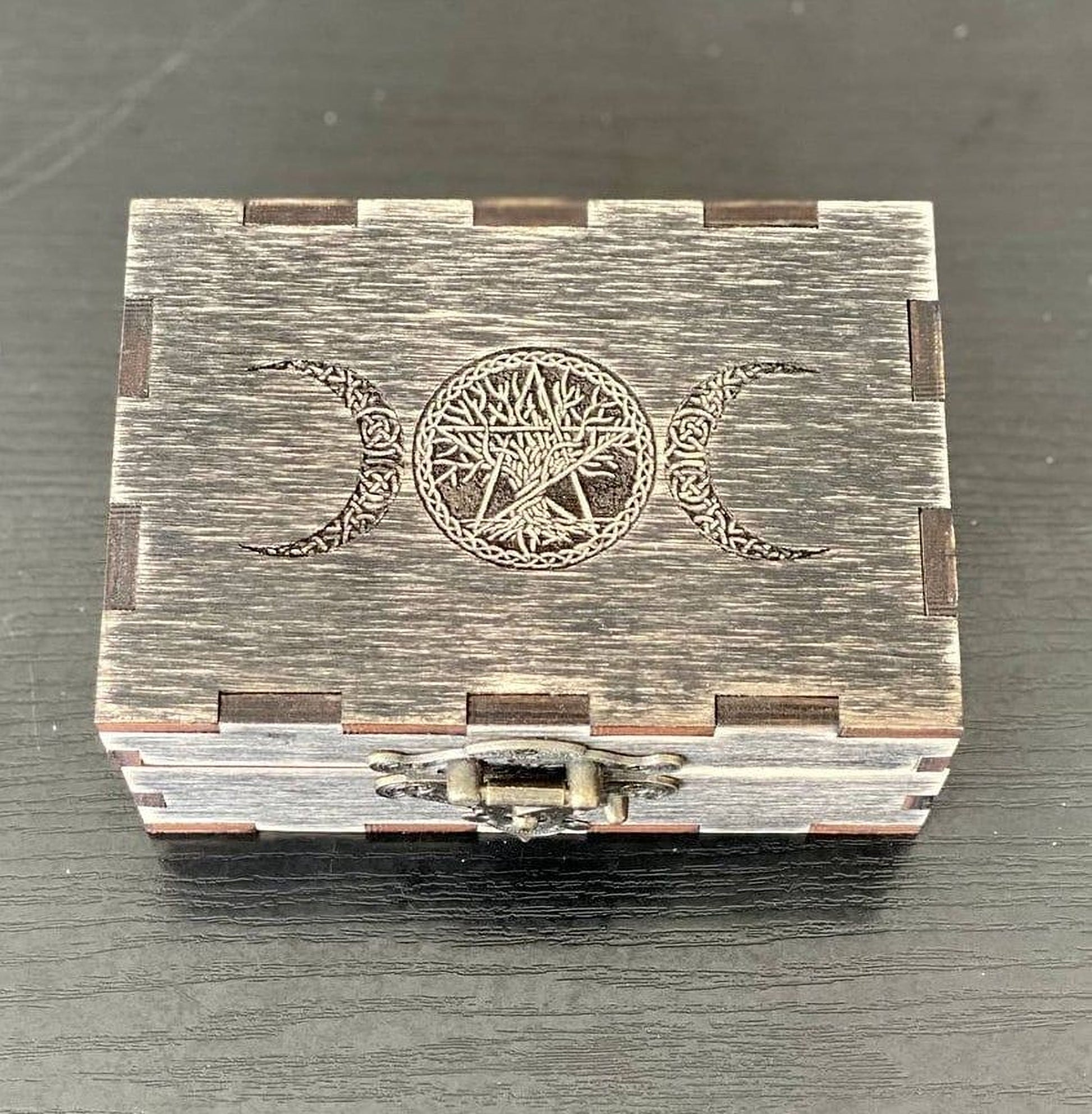 A wooden box with a metal clasp and a carved design on the lid, featuring a tree and crescent moon motif.