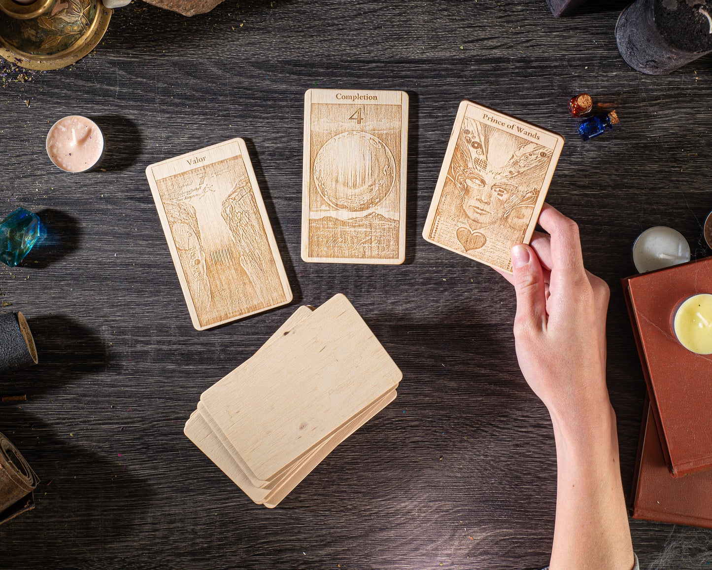 a person's hand holding a wooden card with an intricate design, surrounded by other wooden cards and various objects on a dark wooden surface.