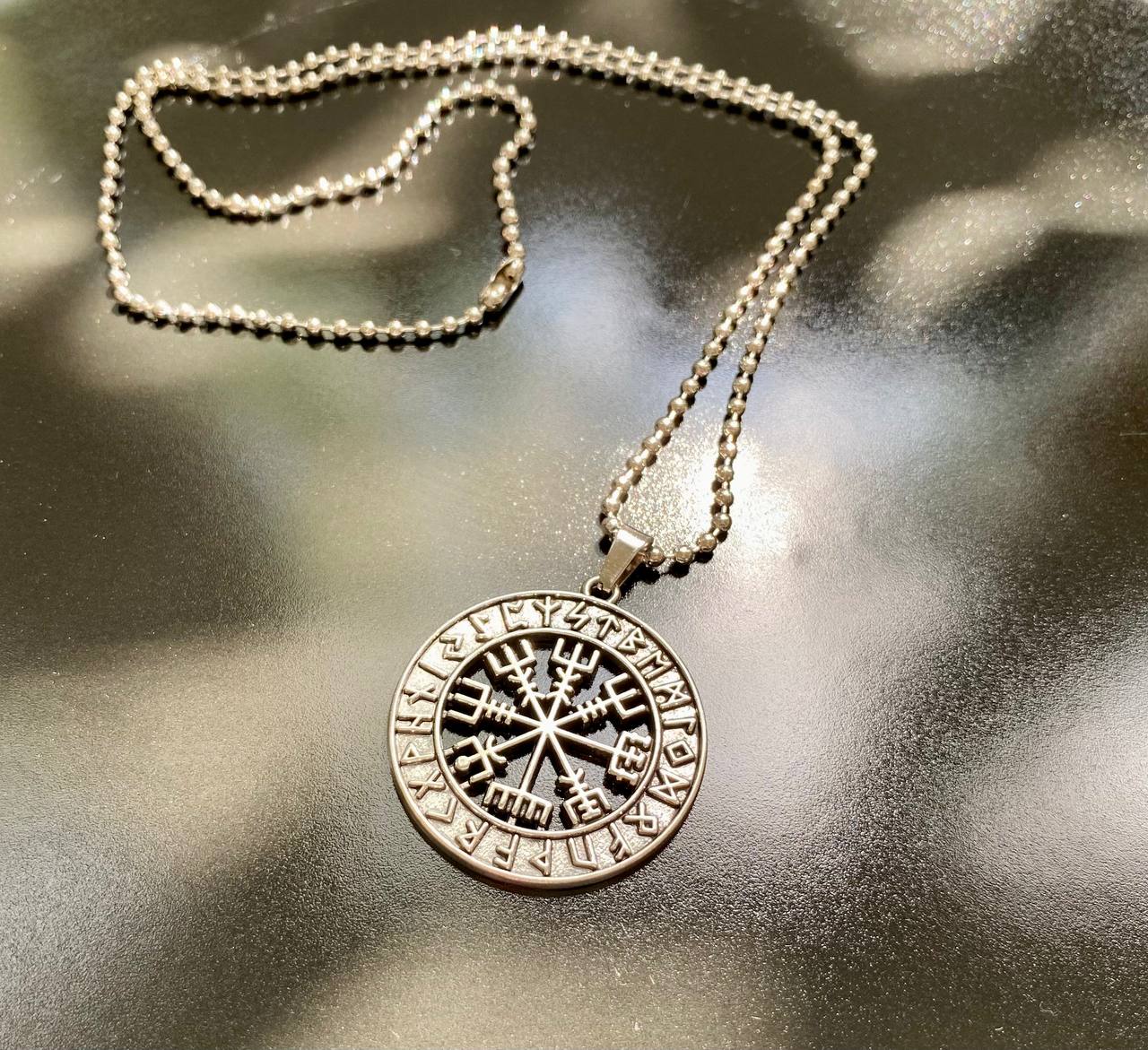 A silver-colored pendant necklace with a circular pendant featuring a compass design and various symbols.