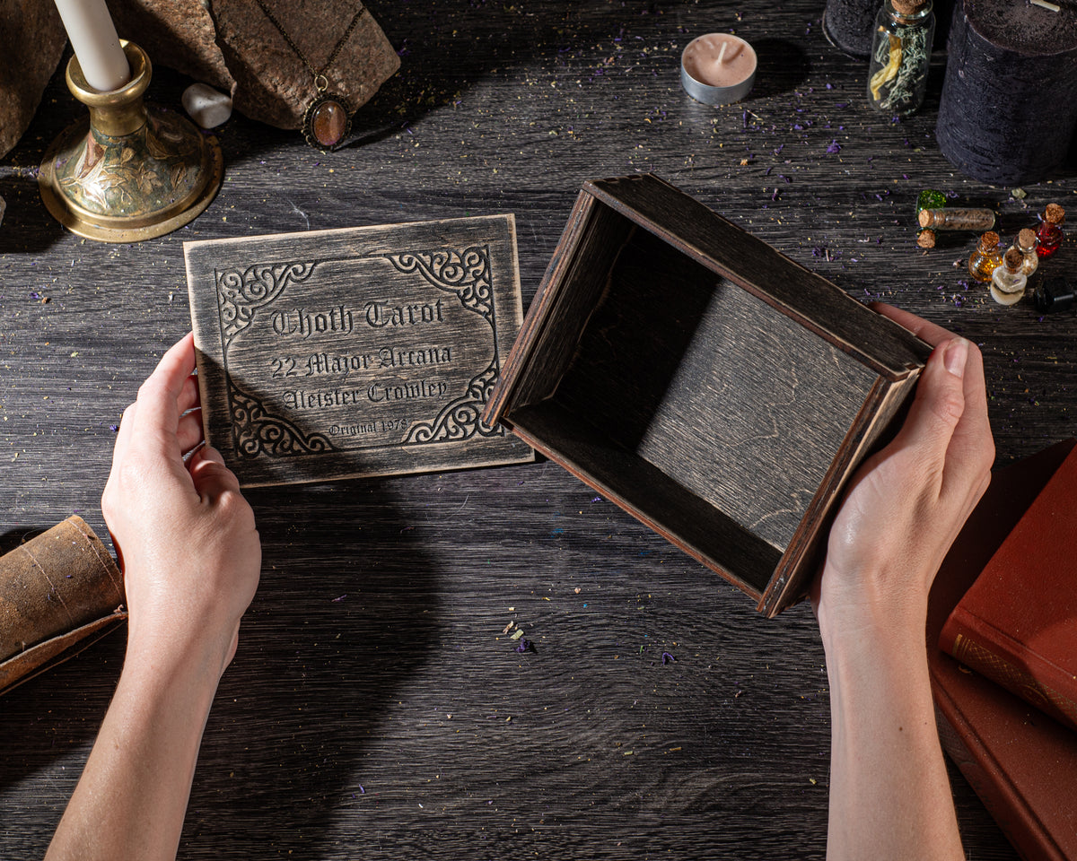 A person's hands holding an ornate wooden box with a decorative label, placed on a wooden surface with various objects and items scattered around.
